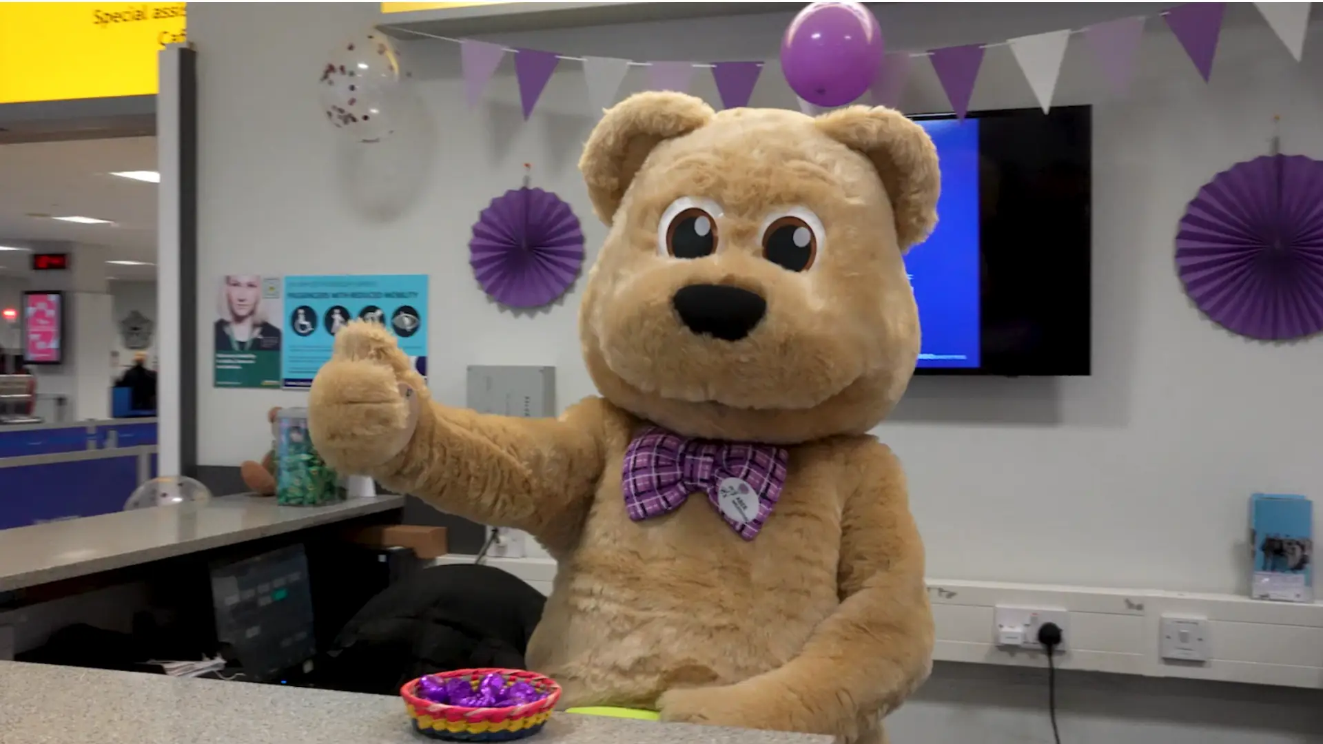 Video Thumbnail for AberNecessities and Aberdeen Airport collab video. A giant teddy mascot behind special services desk